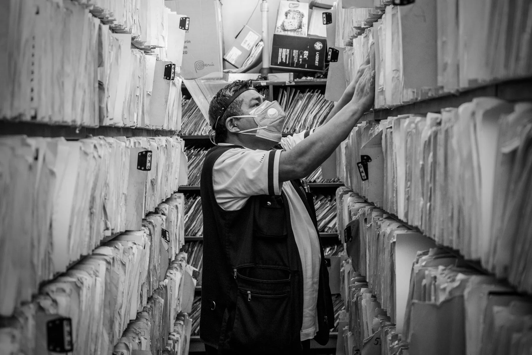A person reaching for a box file among dense archive shelving, in black and white