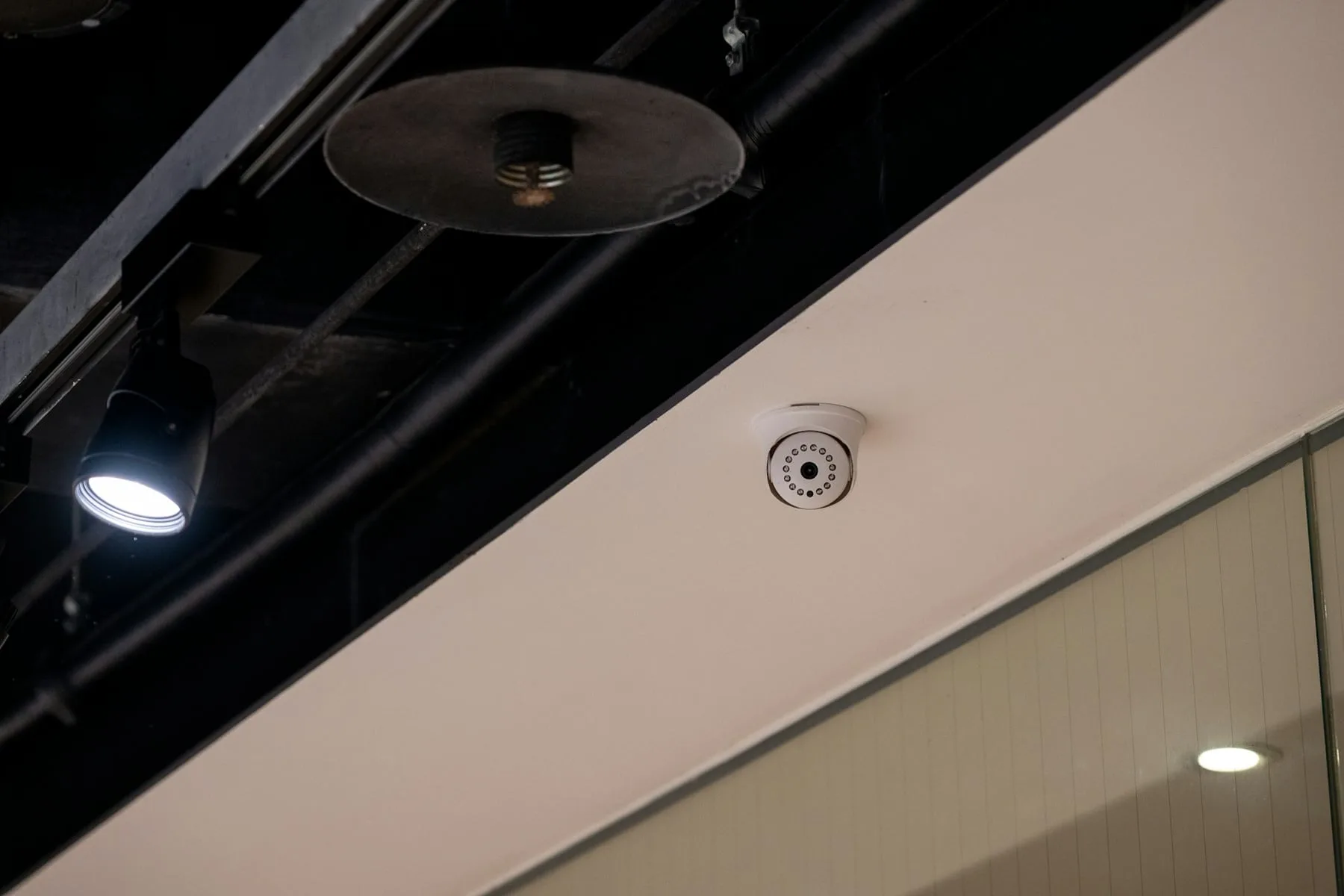 A white dome security camera on a ceiling beside black pipes and a spotlight