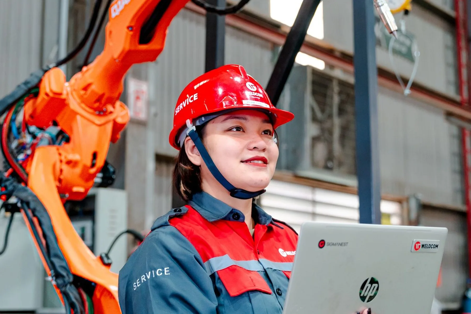An engineer in a hard hat working on a laptop beside an orange robot arm