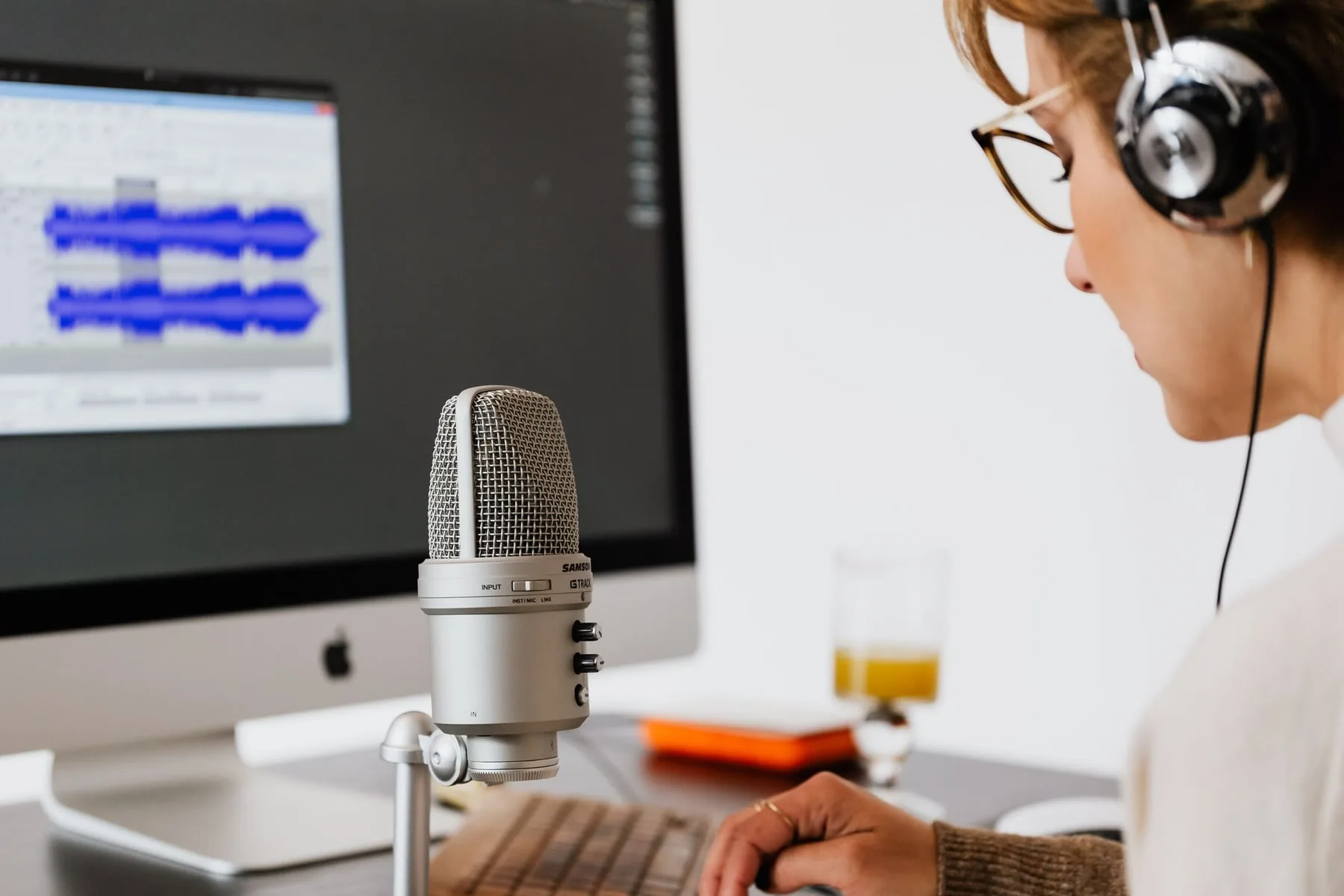 A person speaking into a studio microphone with an audio waveform open on the screen beside them