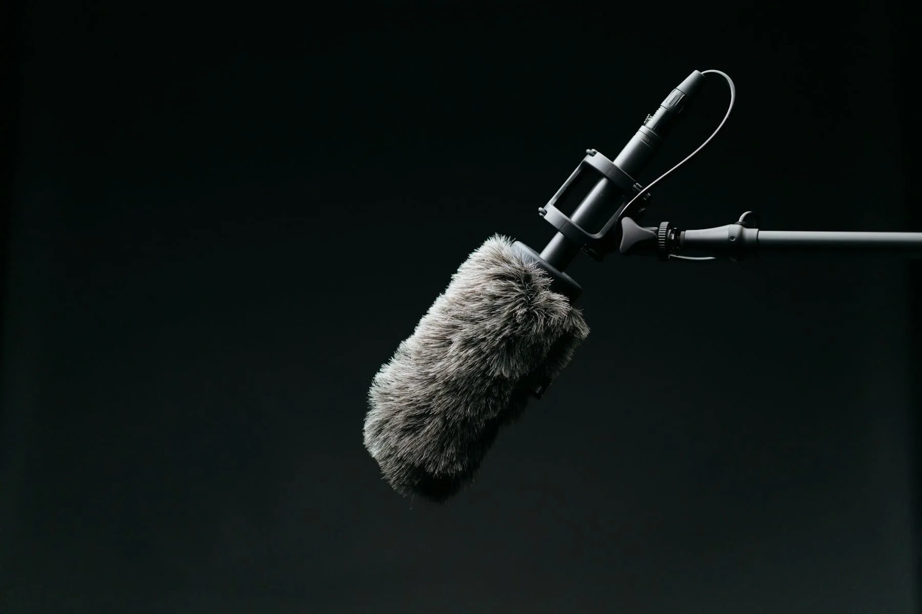 A shotgun microphone with a wind muff on a boom arm, lit against a dark background